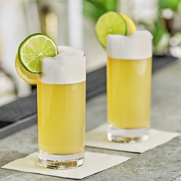 Two glasses of yellow liquid with lime slices on top served in a yellow container with a white lid.