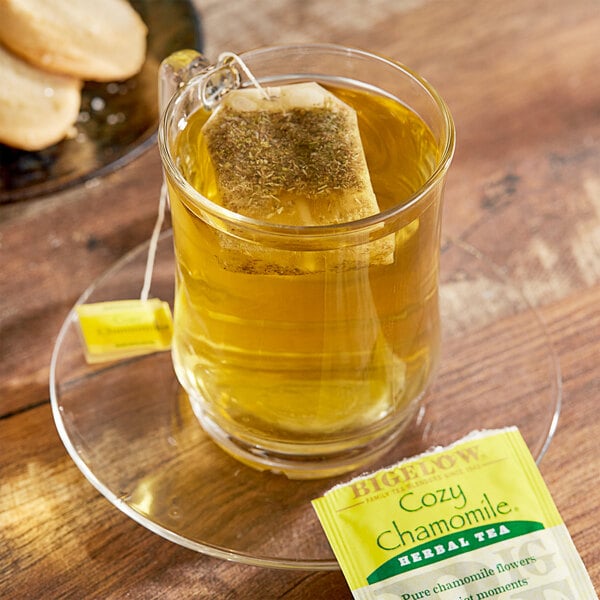 A glass mug of Bigelow Cozy Chamomile tea with a bag of tea on a plate with cookies.