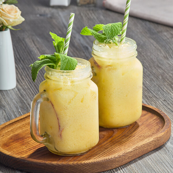 A wooden tray with two glasses of yellow smoothies, a jar of yellow liquid, and mint leaves.