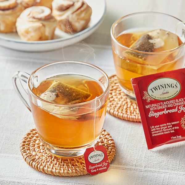 A glass cup of Twinings Gingerbread Joy tea with a tea bag in it, on a coaster with a cinnamon roll.