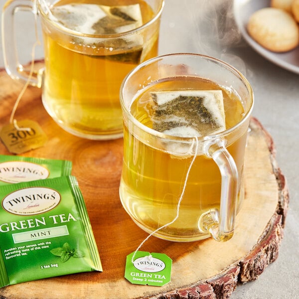 Two mugs of Twinings Green Tea with Mint on a wood table with a tea bag in one mug.