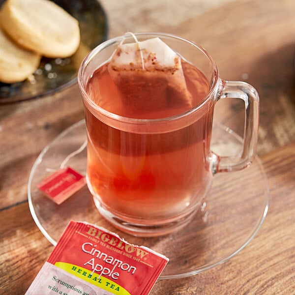 A glass mug of Bigelow Cinnamon Apple Herbal Tea with a tea bag in it on a plate with cookies.