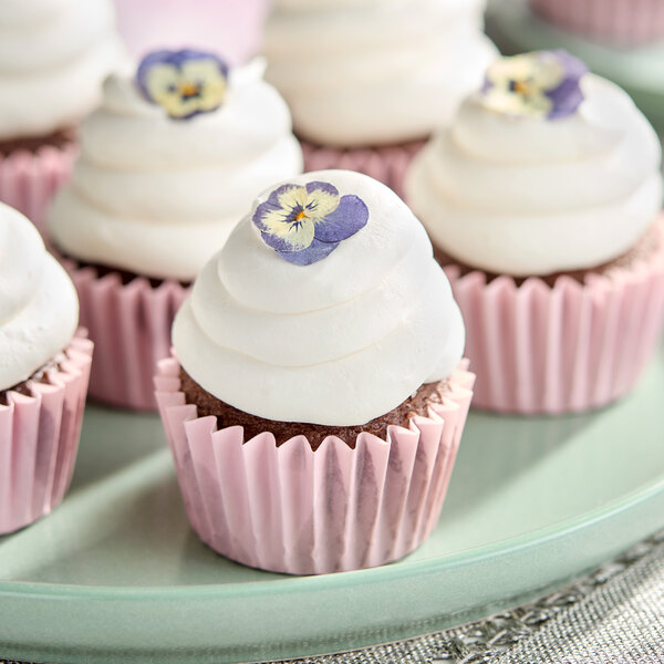 A group of chocolate cupcakes with white frosting and edible purple flowers, each in a pink paper liner, displayed on a plate.