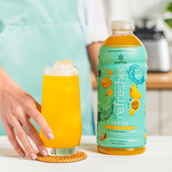 A hand holding a glass of yellow Smartfruit Revive next to a yellow bottle of Smartfruit Revive.