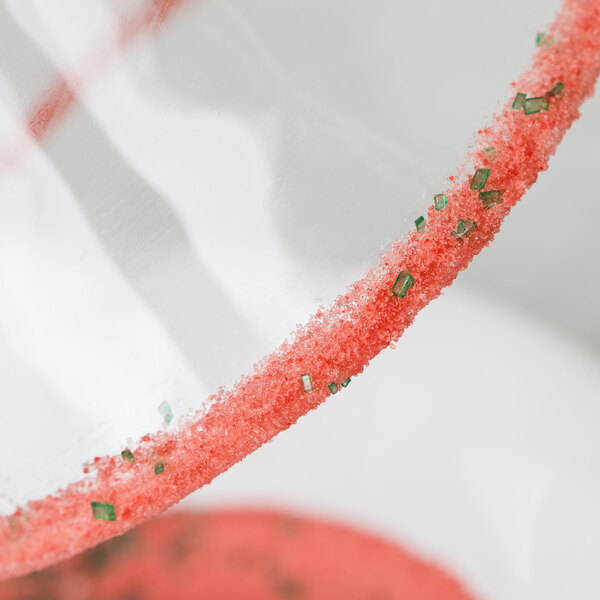 A close up of a glass with a watermelon cocktail rimmed with red and green sugar.