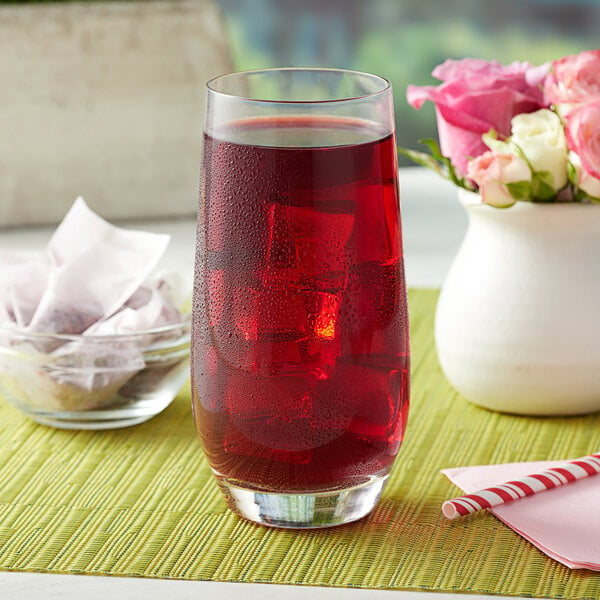 A glass of Bossen Rose Garden fruit tea on ice with a straw on a table with a white vase of flowers.