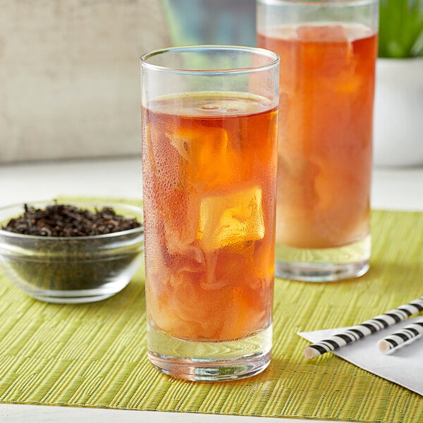 A glass of iced black tea with ice cubes, placed on a green mat next to a bowl of loose leaf black tea and striped straws.