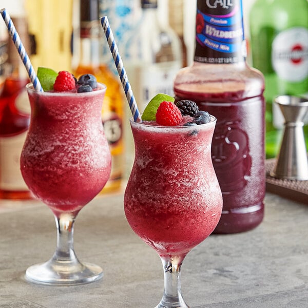 Two glasses of red wild berry drinks garnished with fruit and straws on a bar counter with a bottle of Finest Call Premium Wild Berry Puree.