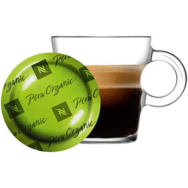 A glass cup of coffee on a green saucer with a Nespresso Peru Organic coffee pod on a green plate with black text.