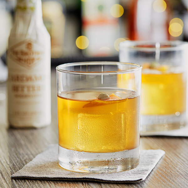 Two glasses of liquid on a table with a bottle of Fee Brothers Cardamom Bitters.
