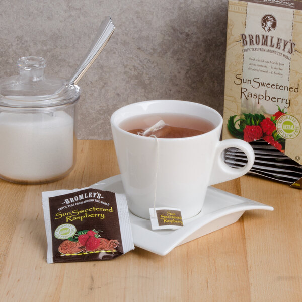 A white mug of Bromley Exotic Sun Sweetened Raspberry Herbal Tea with a tea bag on a table.