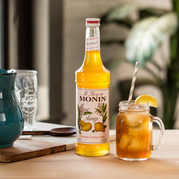 A bottle of Monin mango syrup on a table next to a glass of mango iced tea.