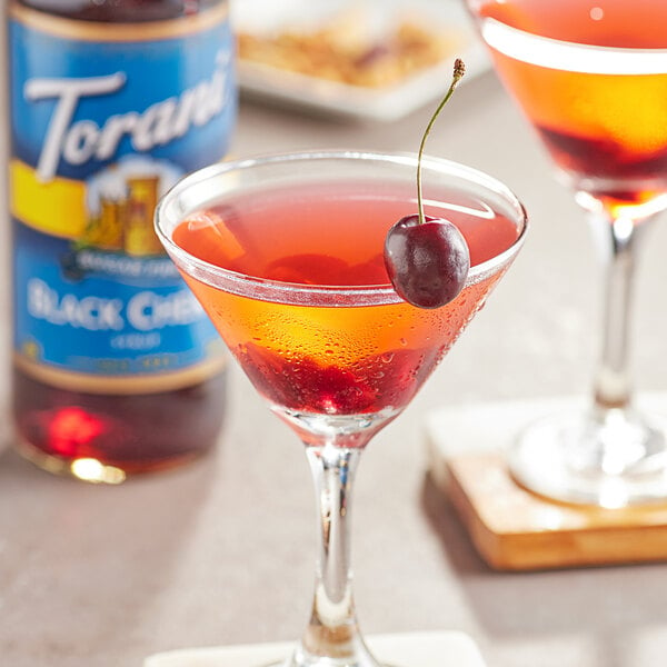 A glass of red liquid with a cherry on top and a bottle of Torani Sugar-Free Black Cherry Flavoring on a table in a cocktail bar.