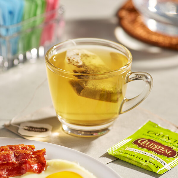 A glass cup of Celestial Seasonings Honey Lemon Ginseng Green Tea with a tea bag in it on a plate of breakfast food.