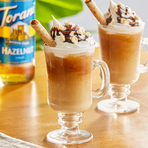 Two glass mugs of coffee with whipped cream and chocolate drizzle on top.