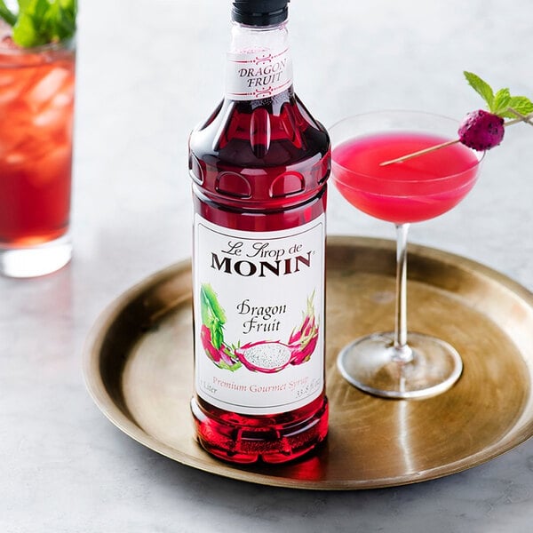 A bottle of Monin dragon fruit syrup next to a glass of pink liquid on a table in a cocktail bar.