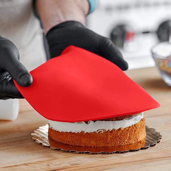 A person wearing black gloves and using a red spatula to cut a cake covered in red Satin Ice fondant.