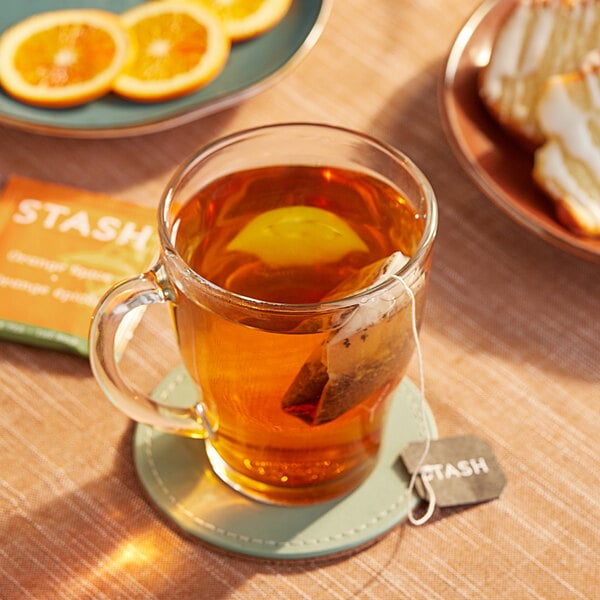 A glass mug of Stash Orange Spice tea with a tea bag in it on a plate with a lemon slice.