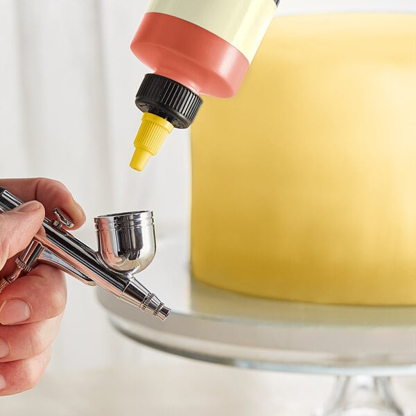 A person using a silver Chefmaster airbrush to spray yellow food coloring on a cake.