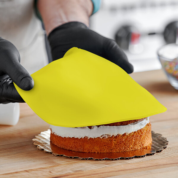 A person wearing black gloves holding a yellow cloth over a cake decorated with yellow Satin Ice rolled fondant.