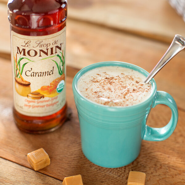 A blue mug with a spoon next to Monin Organic Caramel Flavoring Syrup.