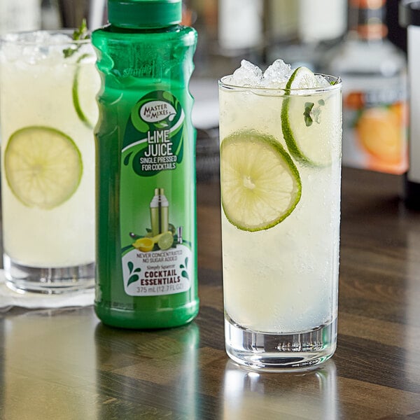 A glass of lime juice next to a green bottle of Master of Mixes Single Pressed Lime Juice.