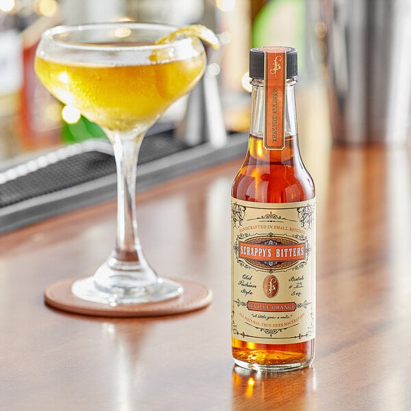 A bottle of Scrappy's Seville Orange bitters on a table next to a glass of yellow liquid.