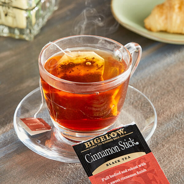 A glass cup of Bigelow cinnamon stick tea on a saucer with a tea bag.