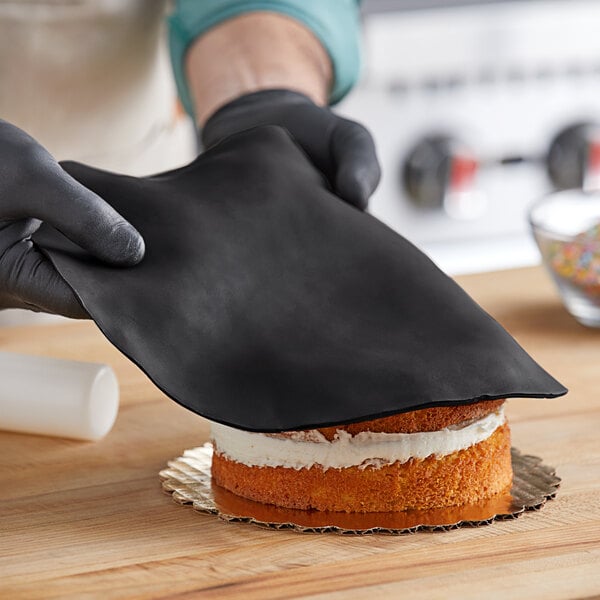 A person wearing gloves rolling out black Satin Ice fondant over a cake.