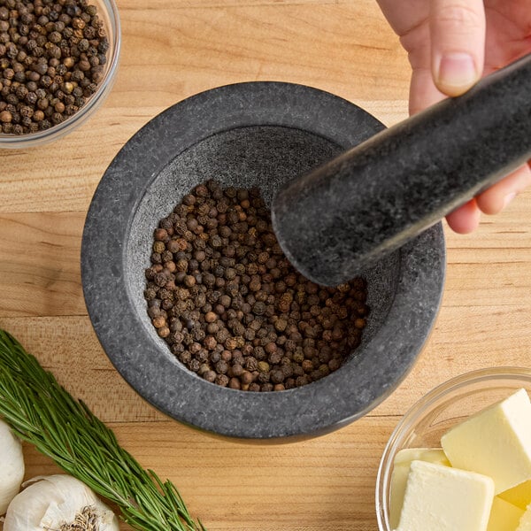 A stone mortar and pestle filled with whole black peppercorns on a wooden surface, surrounded by herbs, garlic, and butter.