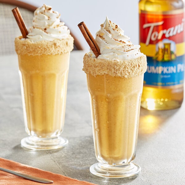 Two glasses of pumpkin pie milkshakes with whipped cream and cinnamon sticks on a table.