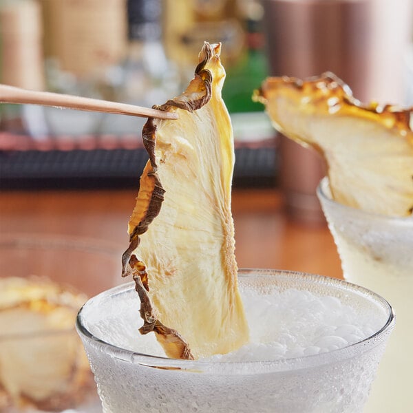 A pair of chopsticks holding a dried pineapple half slice in a glass of liquid.