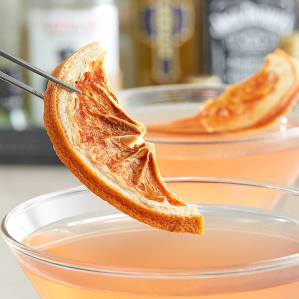 Dried grapefruit half slice held by tweezers over a glass of liquid.