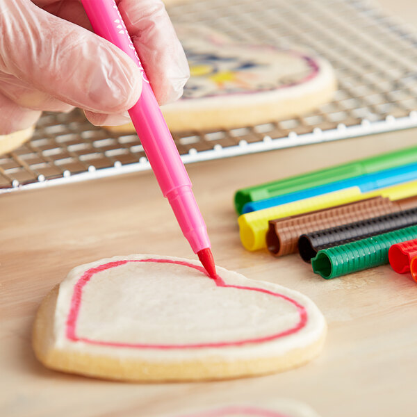 A hand using a pink Chefmaster food decorating pen to draw a heart on a cookie.