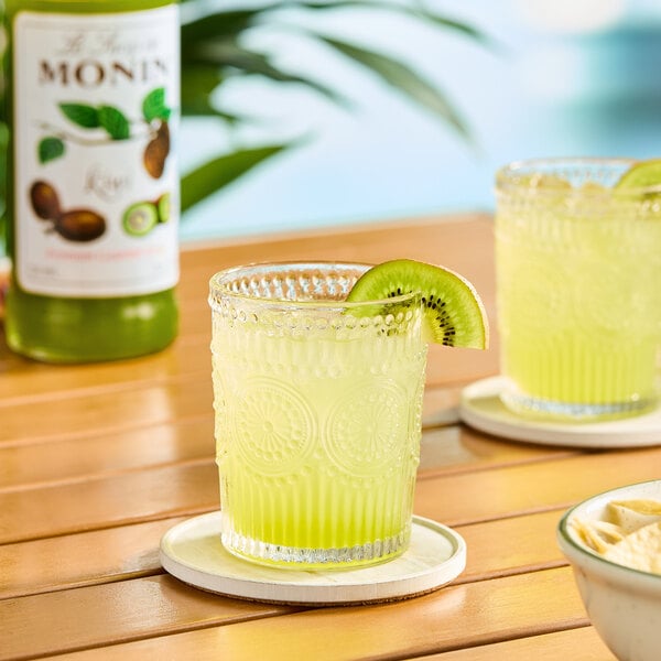 A glass of kiwi-flavored beverage garnished with a slice of kiwi, with a bottle of Monin Premium Kiwi Flavoring Syrup in the background.