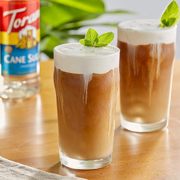 Two glasses of brown liquid with Torani Cane Sugar Sweetener syrup on a table with mint leaves on top.