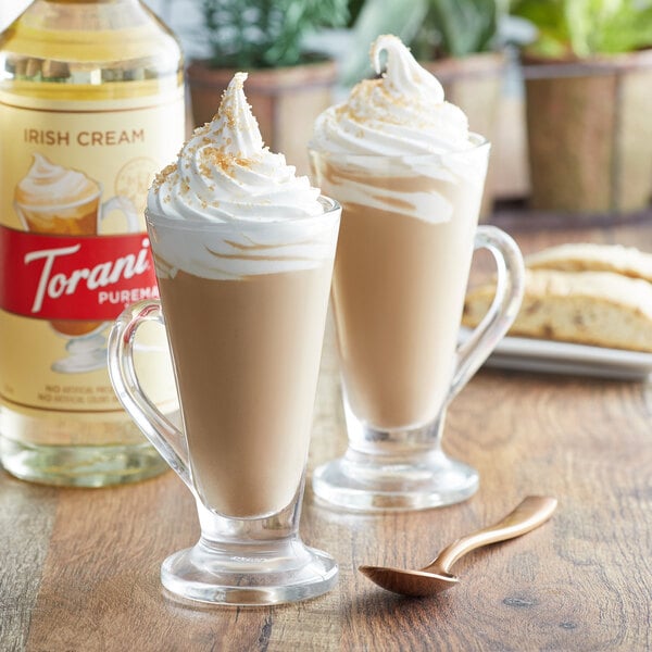 Two glasses of coffee with whipped cream and Torani Irish Cream syrup next to a bottle of Torani Irish Cream syrup.