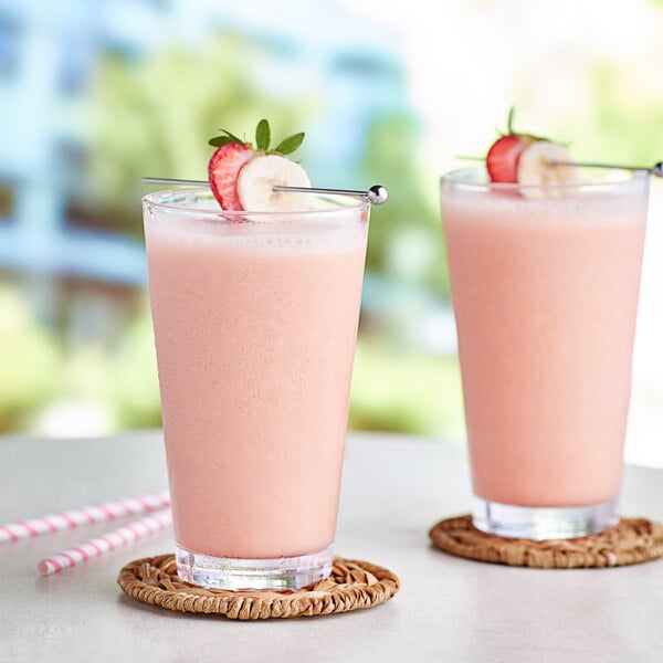 Two glasses of Capora strawberry banana smoothie on a table with fruit and straws.