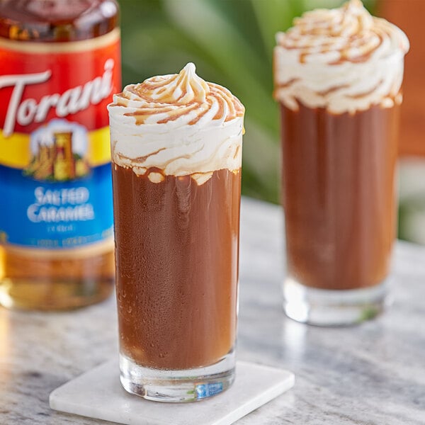 A glass of brown liquid with whipped cream on top next to a bottle of Torani Salted Caramel Flavoring Syrup.