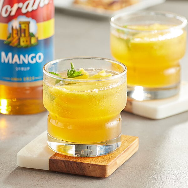 A glass of yellow liquid with a green leaf next to a Torani Mango Flavoring bottle.