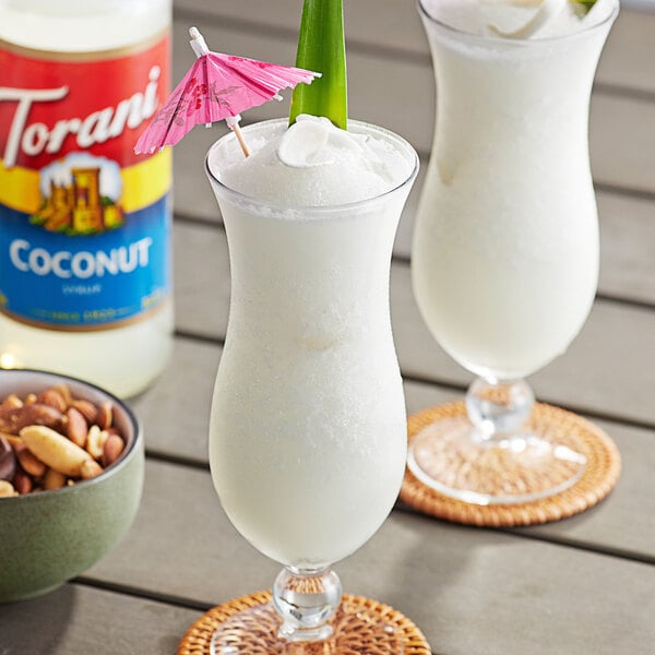 Two white tropical drinks with coconut and pineapple, garnished with nuts and pink umbrellas.