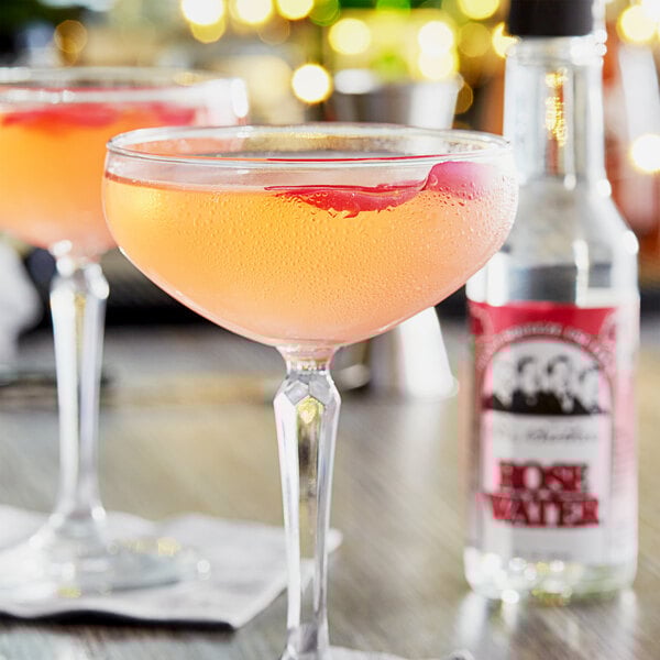 Two glasses of wine on a table with a bottle of Fee Brothers Rose Water.