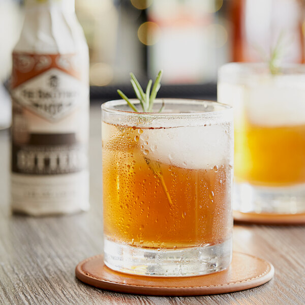 A glass of whiskey and a glass of beer on a table with a bottle of Fee Brothers Whiskey Barrel-Aged Bitters.