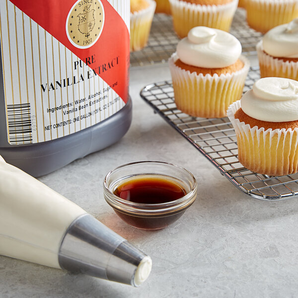 A cupcake with white frosting and a bowl of brown liquid next to a container of Nielsen-Massey Pure Vanilla Extract.