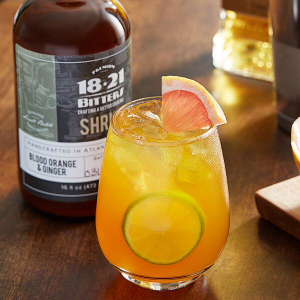 A glass of orange liquid with a slice of lime and a bottle of 18.21 Bitters Blood Orange & Ginger Shrub.