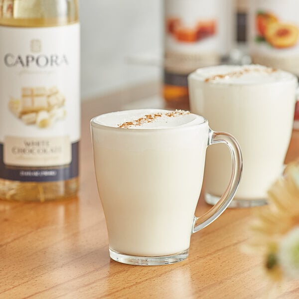 A glass mug of white liquid flavored with Capora White Chocolate syrup next to a bottle of Capora White Chocolate Flavoring Syrup.