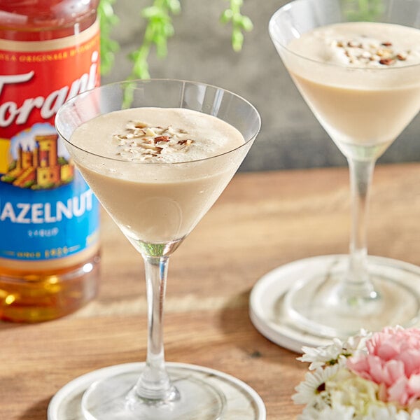 A martini glass filled with brown liquid on a table next to a bottle of Torani Hazelnut Flavoring Syrup.