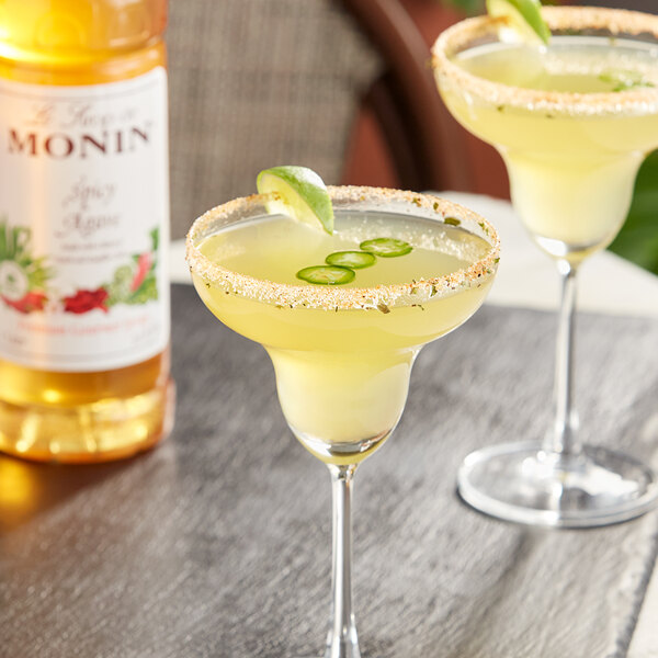 Two glasses of yellow Monin Spicy Agave Syrup margaritas with lime and salt on the rim on a table with a bottle of Monin Spicy Agave Syrup.