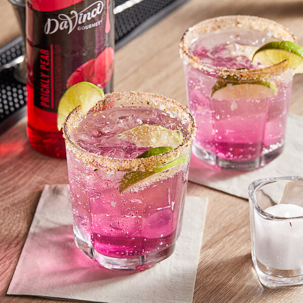 Two glasses of pink DaVinci Gourmet Prickly Pear drinks with limes on a table.