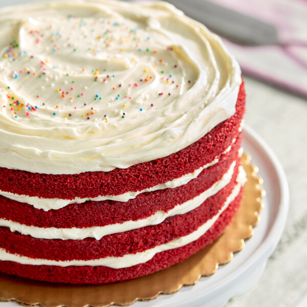 A four-layer red velvet cake with cream cheese frosting and colorful sprinkles on top, displayed on a cake stand.
